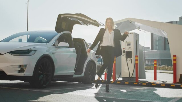 Charming young woman in business suit dancing joyfully outdoors while her luxury electro car charging on EV station. Enjoyment of free time with modern eco friendly transport. Dancing while charging.
