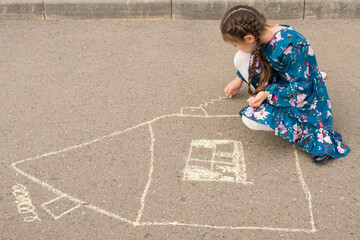 Naklejka premium Junior school girl draws windows and curtains of house with white chalk on asphalt. Girl in flowery dress enjoys drawing house on asphalt, side view