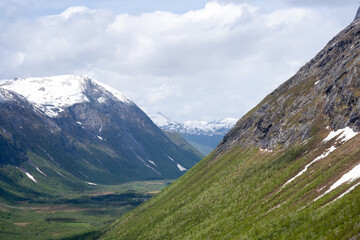 early spring day when there is snow on the mountain and the rocks are gray but above them is a blue sky with white clouds and green grass can be seen at the bottom of the mountain