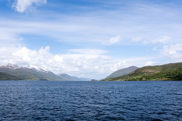 Norway's blue fjord with nearby mountains above which is a beautiful blue sky with puffy white clouds