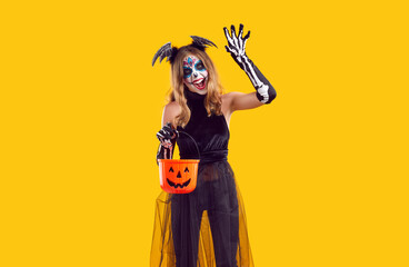 Trick or treat. Preteen girl in creative costume scares, attacks and scratches during Halloween fun. Teenage girl with makeup and Halloween costume is holding candy bucket on orange background.