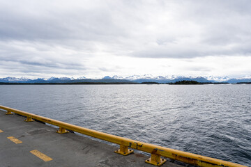 A Norwegian fjord on a harsh overcast day, with dark clans with white snow in the distance and a dramatic sky, but in the foreground gray asphalt with a yellow line and a yellow low metal railing.