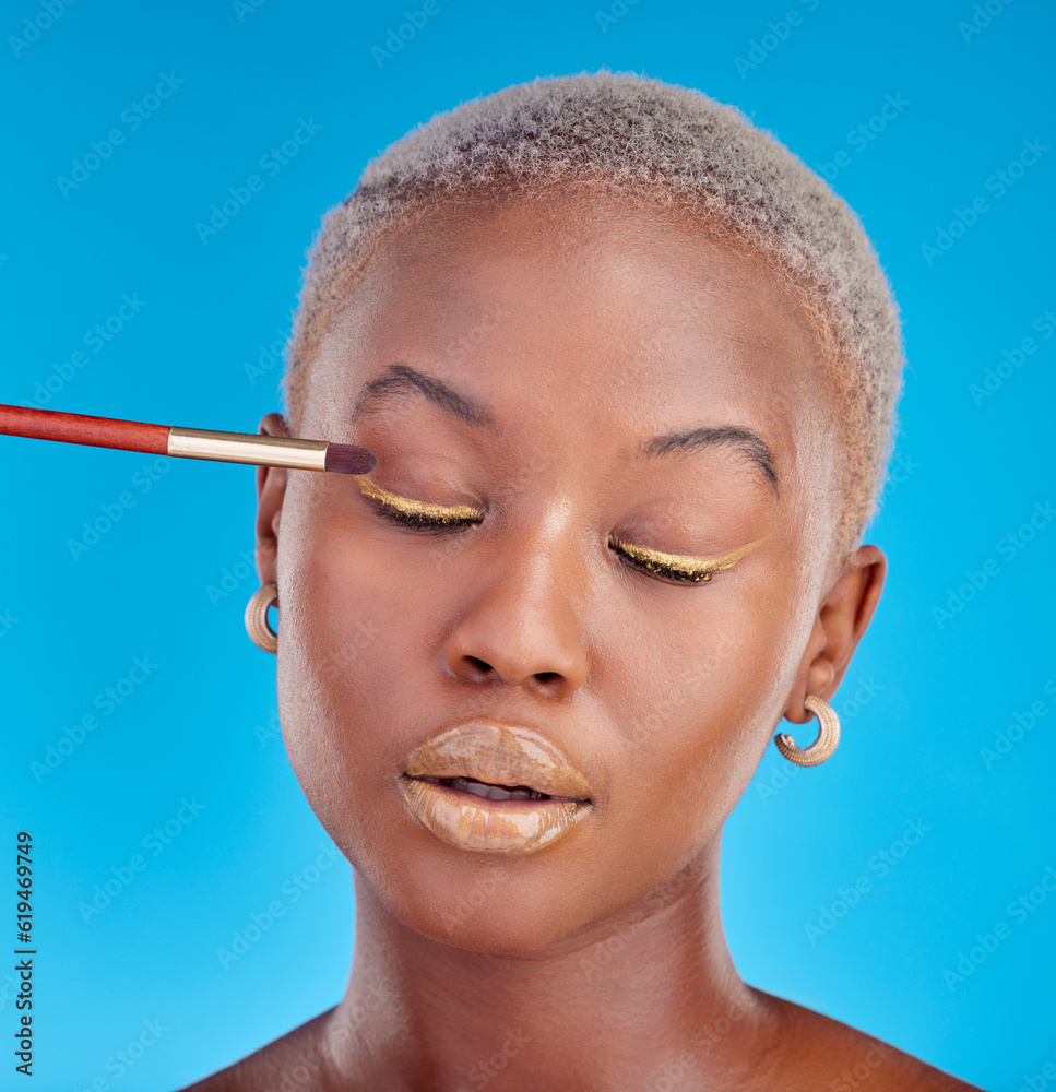 Poster Face, makeup and eyeshadow with a black woman on a blue background in studio to apply a beauty product. Cosmetics, gold color on a brush and a young female model eyes closed for style or glamour