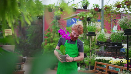 Smiling Young Man with Flower at Horticulture Shop. Redhead Employee in Green Apron in Local Store