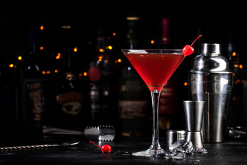 Manhattan alcoholic cocktail drink with bourbon, red vemuth, bitter, ice and cocktail cherry in glass, dark bar counter background, bar tools and bottles