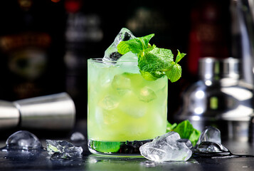 Green Negroni alcoholic cocktail drink with dry gin, aperitif, gentian liqueur, mint and ice cubes. Black bar counter background, bar tools, bottles