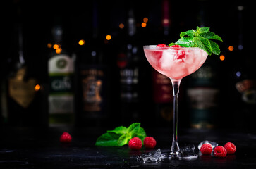 Raspberry daiquiri alcoholic cocktail with white rum, sugar syrup, fruit juice, lime, berry and ice in glass. Black bar counter background, bar tools and bottles