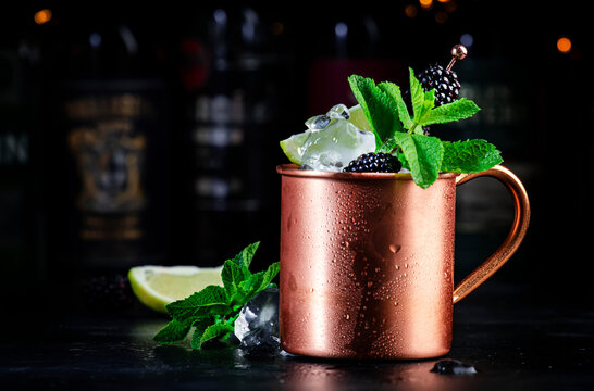 Blackberry Moscow mule alcoholic cocktail drink in copper mug with lime, ice, ginger beer, vodka and mint. Black bar counter background, bar tools, bottles