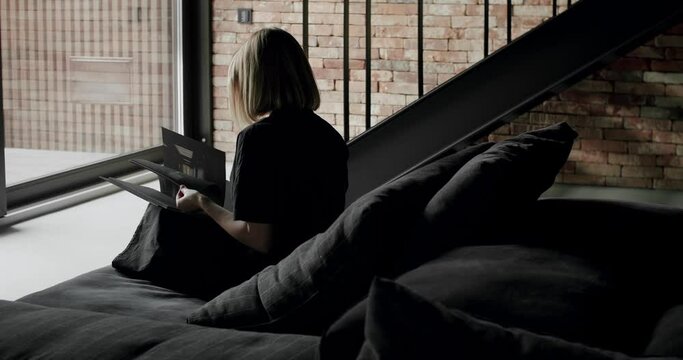 A woman is sitting on the black sofa and reading a book. Modern contemporary Minimalist living room with bricks wall, large sliding windows and black furniture. Fashionable furniture.