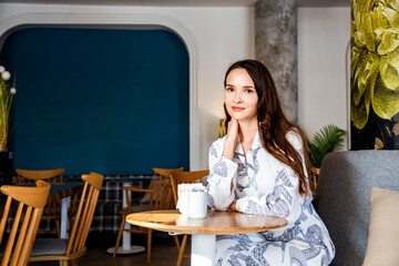 Beautiful young woman with long brown hair sitting in cafe and dreaming about meeting. Brunette with happy face is smiling and waiting. Expectation concept 