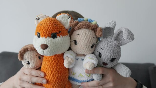 An adorable female craftswoman shoots a video showing off her extraordinary knitted amigurumi soft toys for children. Chanterelle, bunny and dolls in the hands young woman doing small business at home