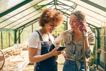 Portrait Of Two Female Friends Working In Greenhouse At Home Looking At Mobile Phone Together