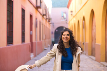 Obraz premium Young and beautiful Hispanic brunette woman with curly hair holds up a hat with her hand while making different expressions and having fun on her city trip in Europe.