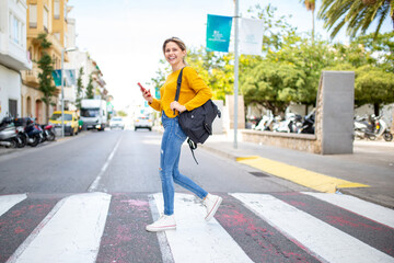 Cheerful young latin woman with handbag and cellphone crossing a street