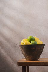Wooden bowl with lemons and limes on wooden bench against concrete wall with shadow. Cozy home interior in minimalist style