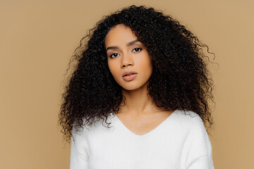 Serious dark-skinned female with frizzy hair, minimal makeup, calmly looks at camera. White jumper,...