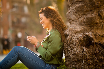 Smiling young latin woman leaning on a tree outside using mobile phone