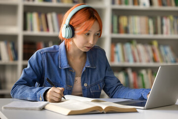 Serious hipster red haired student girl in big headphones studying in college library, writing notes at laptop, open book, listening to music for focus, watching learning class, webinar, lesson
