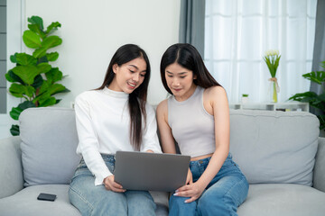 Two young asian women shopping online shopping on laptop while sitting on sofa together in living room.