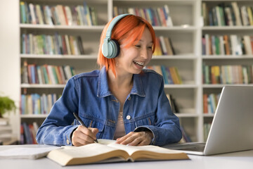Happy joyful college girl with red hair and teeth brackets using headphones and laptop for studying in campus library, writing notes, watching online educational content, laughing