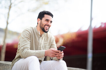 Cheerful young middle eastern guy listening to music from phone sitting outside