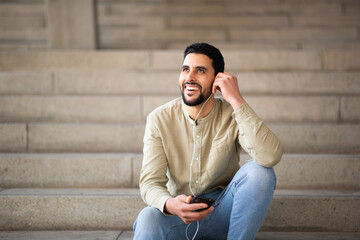 Happy young arabic man listening to music with cellphone sitting on steps