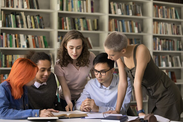 College classmates reading textbook in campus library together, working on group research study. University students standing, sitting at table with bookshelves in background, reviewing book