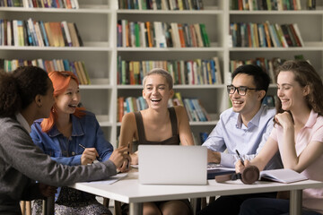 Multiethnic team of happy fresh students cooperating on class project together, enjoying teamwork in campus library, writing notes at laptop, talking, laughing, discussing college education