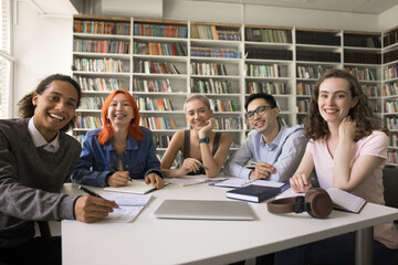 Happy diverse college fresh students posing for group portrait in campus library, sitting at table with bookshelves in background, writing notes, studying books, looking at camera, smiling - Powered by Adobe