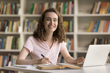 Happy beautiful student girl working on article in college library, writing essay, sitting at table with open books, laptop, looking at camera, smiling, laughing, promoting university education