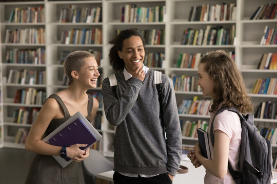Cheerful diverse student girls and guy talking in college library together, laughing, discussing education, enjoying friendship. Happy high school classmates with backpacks having fun in campus