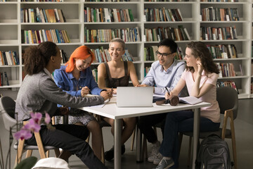 Happy trendy classmates doing school homework in library together, sitting at table, laughing,...