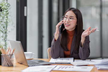 Cheerful business Asian woman freelancer wear glasses making telephone call share good news about project working in office workplace, business finance concept.