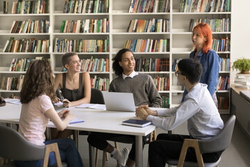 Teen red haired student girl standing at library table, speaking to diverse group of friends...