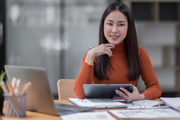 Fototapeta premium Attractive smiling young asian business woman wear glasses work at home office, Asian woman working on laptop computer holding tablet.business finance concept.