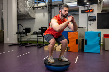 Fit athletic man performing exercise on gymnastic hemisphere bosu ball in gym.