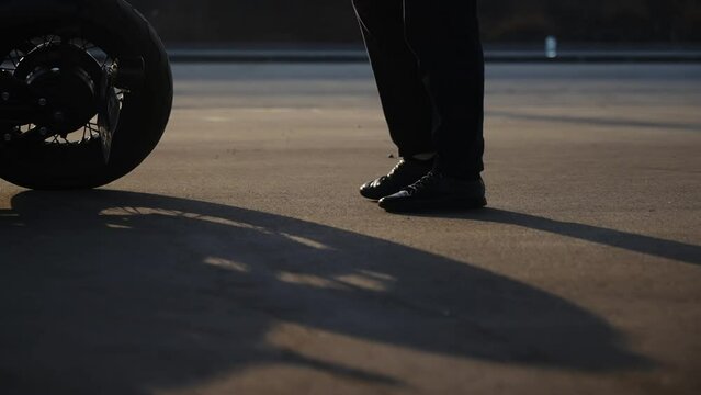 Men's feet in black leather boots fit the part of the motorcycle. A man in black biker pants walks along an asphalt road in a parking lot. The dynamic camera moves parallel to the legs. Black and