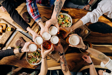 Friends cheering beer glasses on wooden table covered with delicious food - Top view of people...
