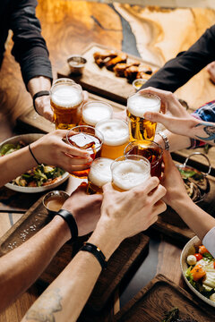 Friends cheering beer glasses on wooden table covered with delicious food - Top view of people having dinner party at bar restaurant - Food and beverage lifestyle concept