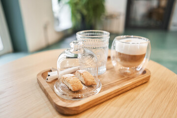 Cup of coffee cappuccino with cantuccini and a glass of water on a wooden tray. Coffee with milk, a cookie and a glass with water on a glass table.