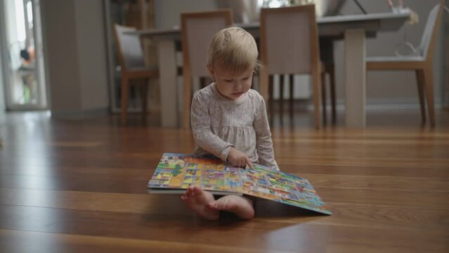 Little baby girl with a book. The child looks at a book with different pictures at home. Happy child reading a book. 2 year old toddler baby infant.