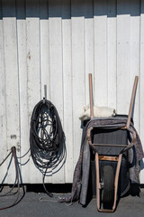 a wheelbarrow propped up against a white barn wall, a fire extinguisher and a bundle of black wire...