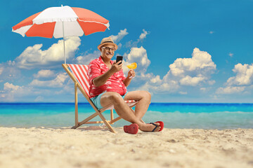 Man at the beach sitting on a deck chair by the sea with a cocktail and using a mobile phone