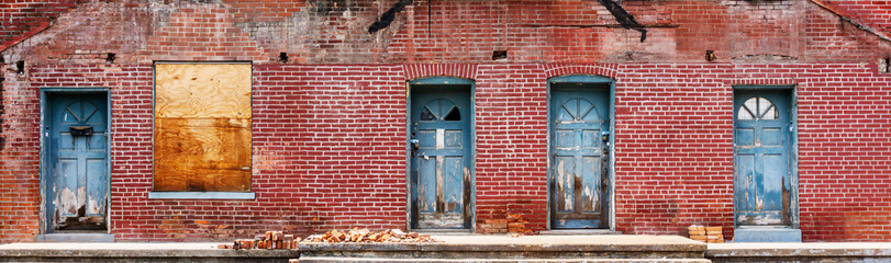 Fototapeta na wymiar Distressed brick building front with four blue doors and a boarded-up window background banner 