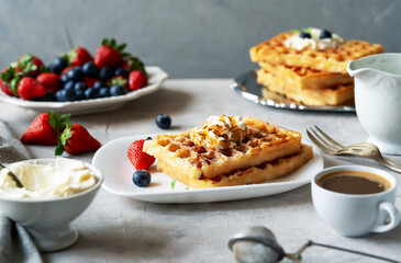 Belgian waffles with berries on white plate, blueberries, strawberries, strainer, dairy cream, cup of coffee on concrete background