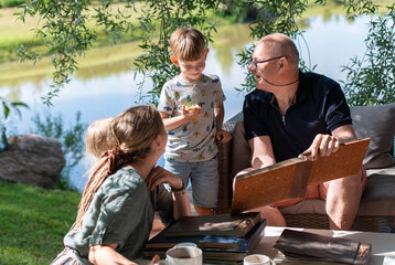 Grandpa with his grandchildren looking at old photo albums on outdoor terrace on a sunny summer day