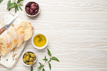 Sliced fresh ciabatta on cutting kitchen board, green and brown olives, olive oil with rosemary, olive tree branches, white wooden rustic background top view copy space