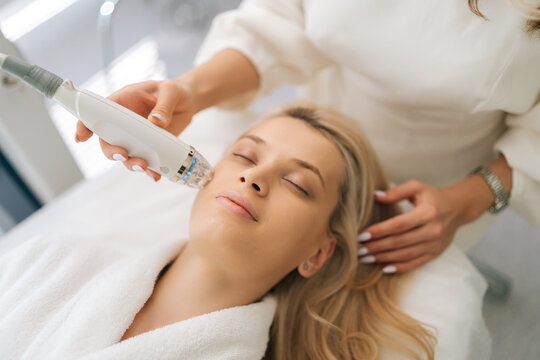 Close-up high-angle view face of blonde woman client lying with closed eyes and getting stimulating beauty facial treatment during rf-lifting procedure. Concept of anti-age rejuvenation skincare.