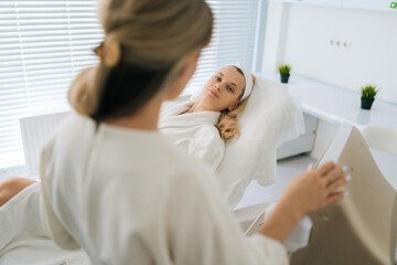 Back view of unrecognizable cosmetologist consulting female client lying on medical couch before face smas lifting procedure in clinic of aesthetic medicine. Concept of anti-age rejuvenation skincare.
