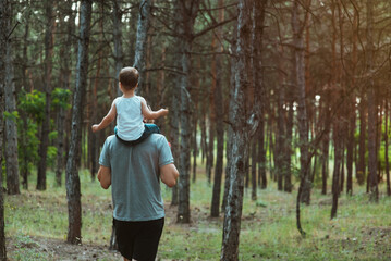 father's day. Happy European-looking father walks with his young cute two-year-old son on his shoulders in the woods